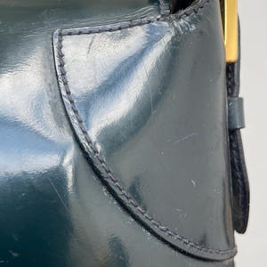 Puede incluir: Un bolso de mano de charol en color verde bosque oscuro con un acabado liso y brillante, con costuras visibles a lo largo de los paneles curvos. Un detalle met&aacute;lico rectangular en tono dorado se encuentra en el lateral, cerca de la correa.
