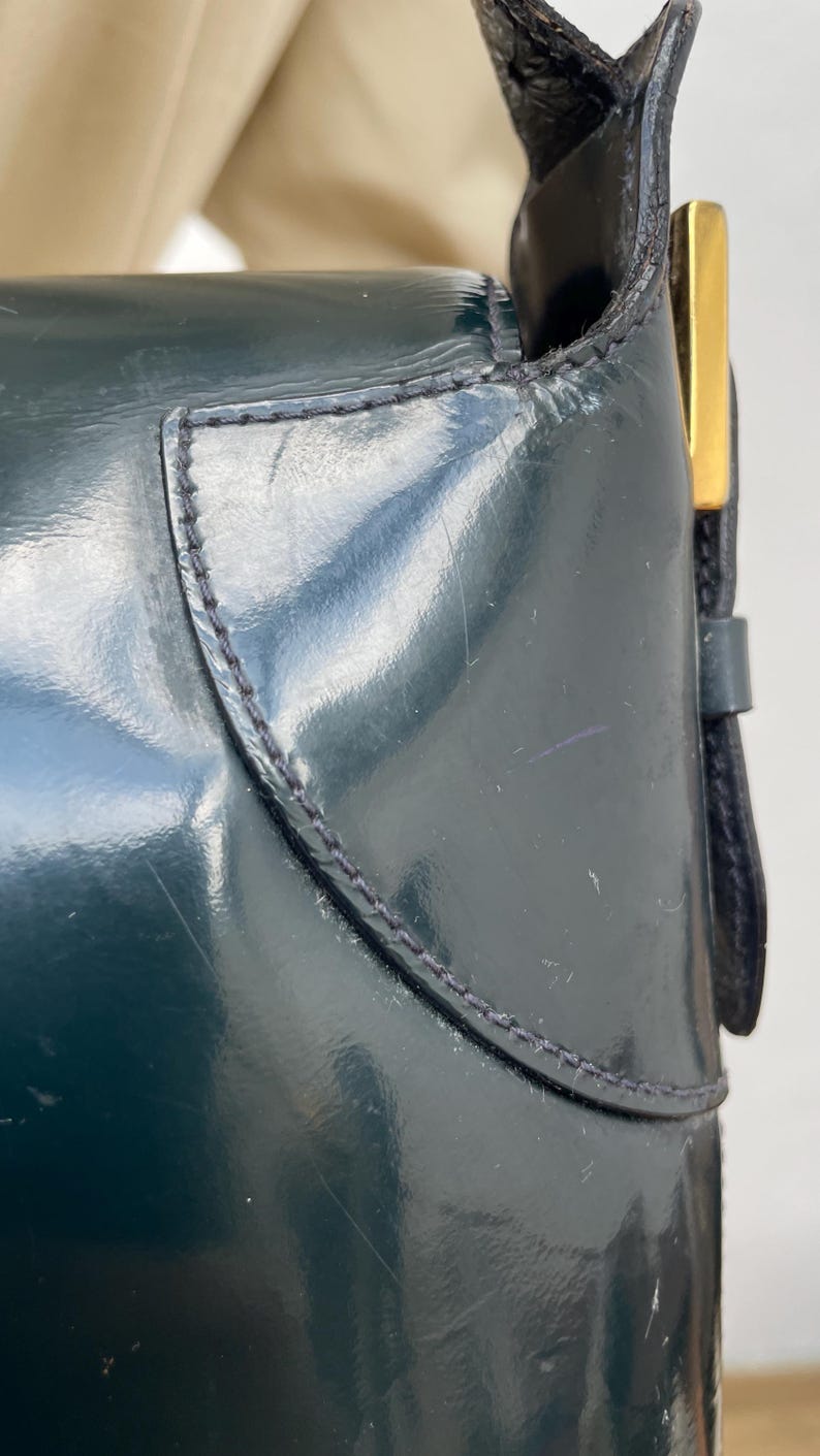 Puede incluir: Un bolso de mano de charol en color verde bosque oscuro con un acabado liso y brillante, con costuras visibles a lo largo de los paneles curvos. Un detalle met&aacute;lico rectangular en tono dorado se encuentra en el lateral, cerca de la correa.