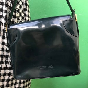Puede incluir: Un bolso de hombro de charol azul marino oscuro con acabado brillante, que presenta un cierre de solapa y la palabra VALENTINO grabada en letras may&uacute;sculas en la parte frontal. El bolso incluye una correa a juego y detalles met&aacute;licos dorados.