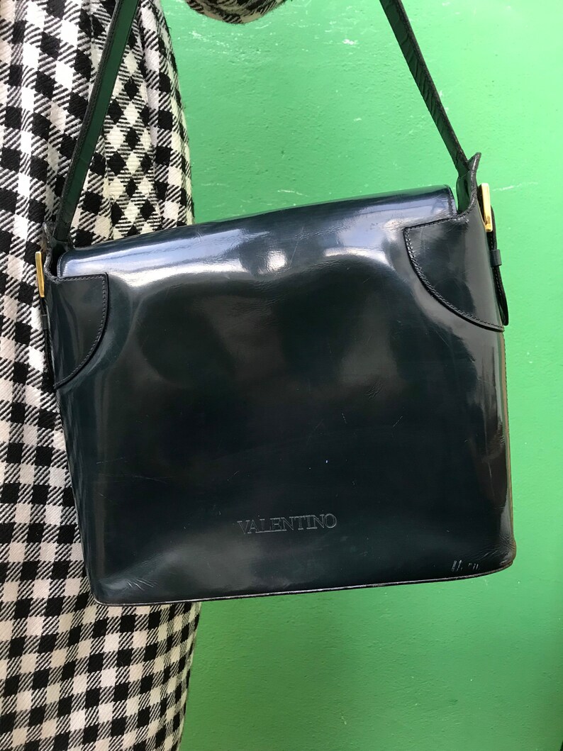 Puede incluir: Un bolso de hombro de charol azul marino oscuro con acabado brillante, que presenta un cierre de solapa y la palabra VALENTINO grabada en letras may&uacute;sculas en la parte frontal. El bolso incluye una correa a juego y detalles met&aacute;licos dorados.