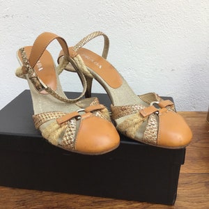 May include: A pair of Prada high-heeled sandals. The shoes feature a tan leather ankle strap and toe cap, with a woven raffia and snakeskin pattern on the sides. The heels are slender and the shoes are set on a black box.