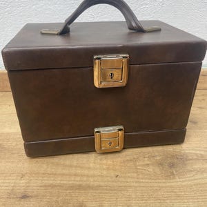 Op de afbeelding: Een bruine vintage reiskoffer met een gebogen handvat en twee goudkleurige sluitingen. De koffer is rechthoekig en lijkt van leer of een vergelijkbaar materiaal te zijn. De sluitingen hebben sleutelgaten, wat suggereert dat de koffer op slot kan.