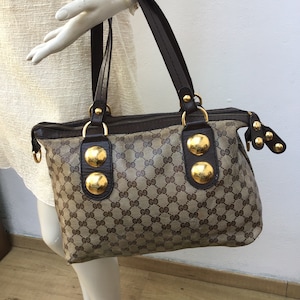 Peut inclure: Sac à main marron et beige avec un motif de logo répété. Le sac a des anses marron foncé et des éléments métalliques dorés, dont de gros clous ronds. Le sac est présenté sur un mannequin.