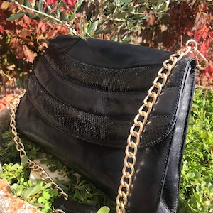 Op de afbeelding: Zwarte leren schoudertas met een gouden kettingriem. De tas heeft een klepsluiting met een gestructureerd, slangachtig ontwerp. De tas ligt op een bed van groen gebladerte, met een achtergrond van rode en groene bladeren.