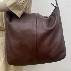 May include: A large, brown leather shoulder bag with a long strap. The bag has a soft, slouchy design and a simple, classic style. The leather appears to have a textured finish. The bag is shown hanging from a shoulder.