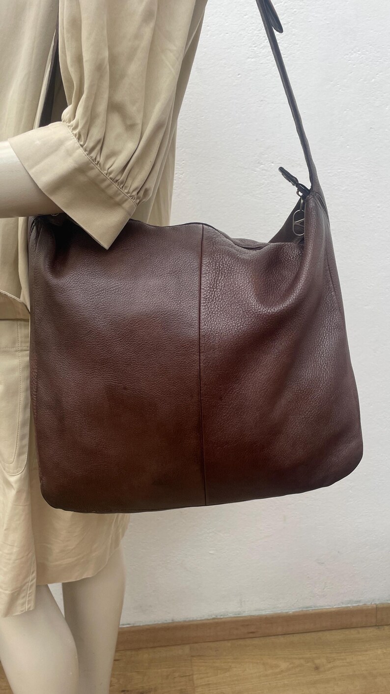 May include: A large, brown leather shoulder bag with a long strap. The bag has a soft, slouchy design and a simple, classic style. The leather appears to have a textured finish. The bag is shown hanging from a shoulder.