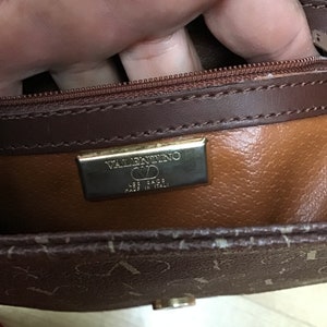 K&ouml;nnte beinhalten: Eine braune Ledertasche mit einem goldenen Rei&szlig;verschluss und einem goldenen Schild mit dem Text "VALENTINO  LES SACS  MADE IN ITALY".