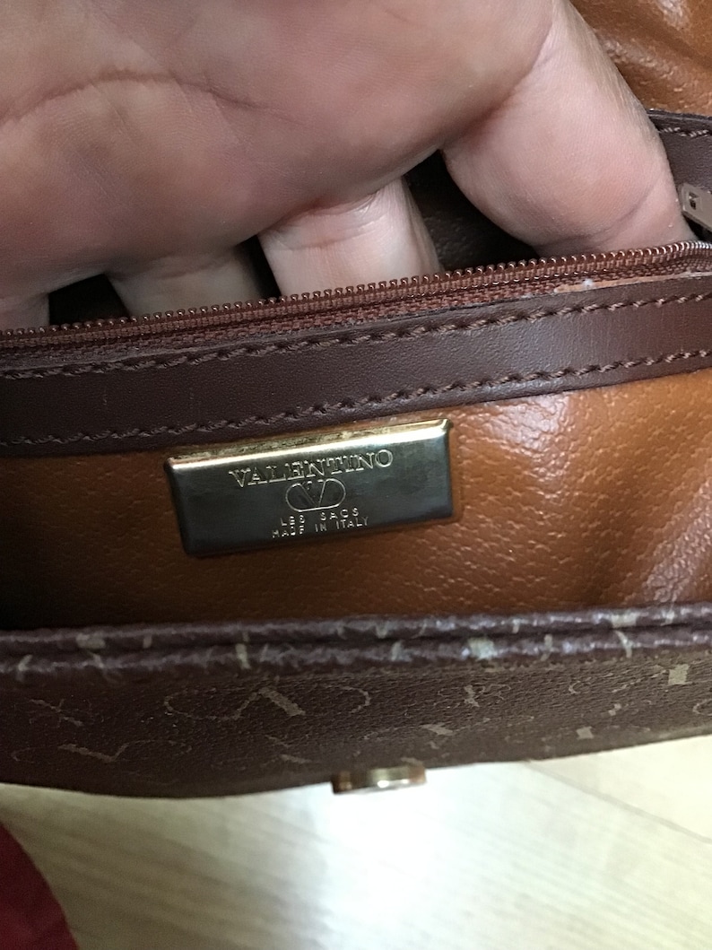 K&ouml;nnte beinhalten: Eine braune Ledertasche mit einem goldenen Rei&szlig;verschluss und einem goldenen Schild mit dem Text "VALENTINO  LES SACS  MADE IN ITALY".