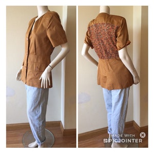 May include: A tan short-sleeved blazer with a decorative patterned back panel. The blazer has a V-neck, button closure, and two front pockets. The mannequin is also wearing light blue trousers.
