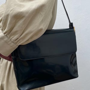 Puede incluir: Un bolso de hombro de color azul marino oscuro con un acabado brillante tipo charol. El bolso presenta una forma rectangular con cierre de solapa y una correa de hombro &uacute;nica sujeta con herrajes met&aacute;licos dorados en el lateral.