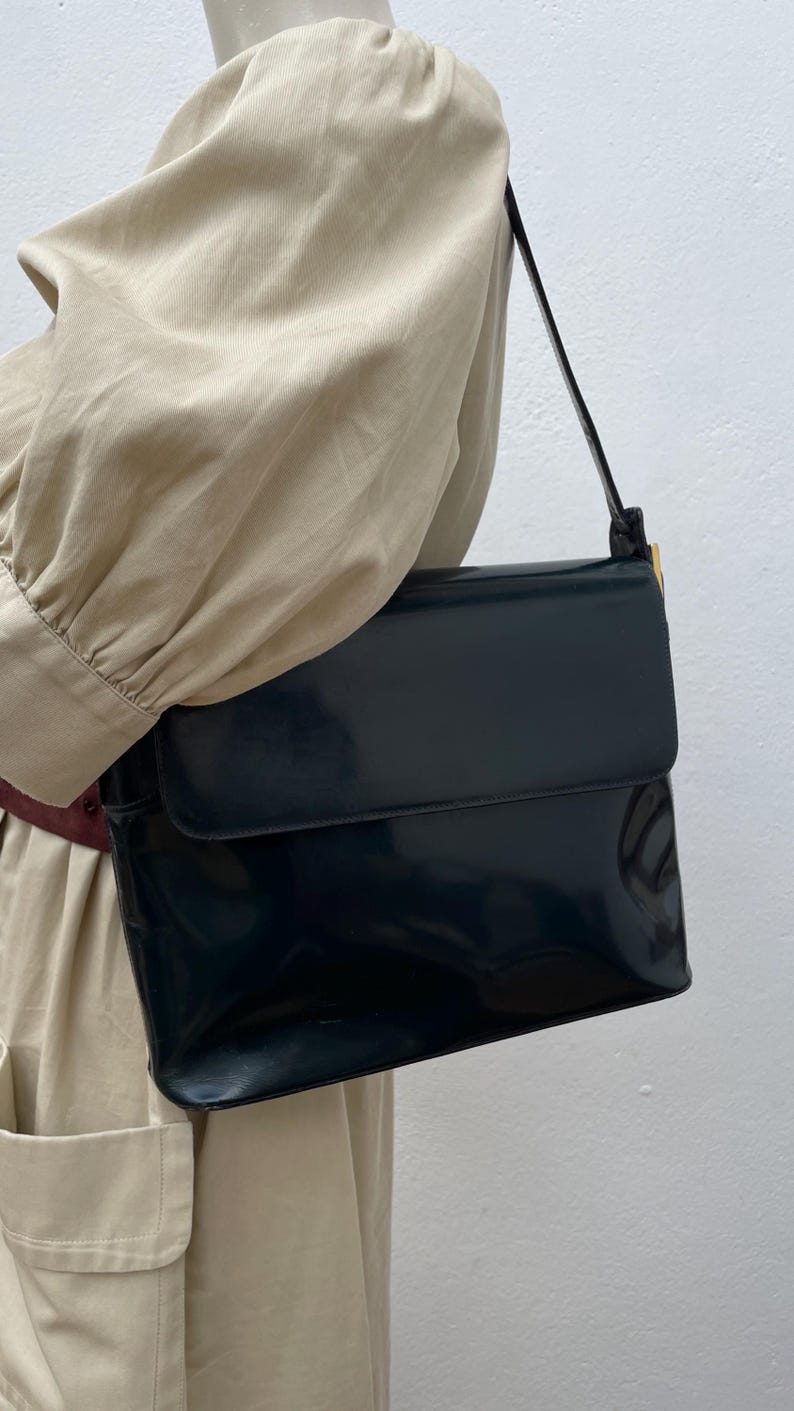 Puede incluir: Un bolso de hombro de color azul marino oscuro con un acabado brillante tipo charol. El bolso presenta una forma rectangular con cierre de solapa y una correa de hombro &uacute;nica sujeta con herrajes met&aacute;licos dorados en el lateral.