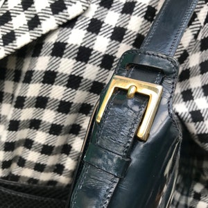 Puede incluir: Una correa de bolso de charol verde oscuro con una hebilla de metal dorado pulido. La correa est&aacute; hecha de un material liso y brillante con textura visible y se encuentra sobre una tela con estampado de pata de gallo en blanco y negro.