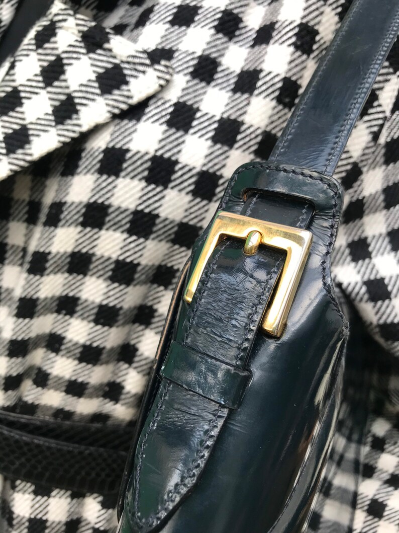 Puede incluir: Una correa de bolso de charol verde oscuro con una hebilla de metal dorado pulido. La correa est&aacute; hecha de un material liso y brillante con textura visible y se encuentra sobre una tela con estampado de pata de gallo en blanco y negro.