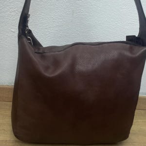 May include: A brown leather shoulder bag with a long strap. The bag is rectangular with a rounded top and a smooth, textured surface. The strap is dark brown and attached to the bag with metal hardware. The bag is sitting on a wooden surface.