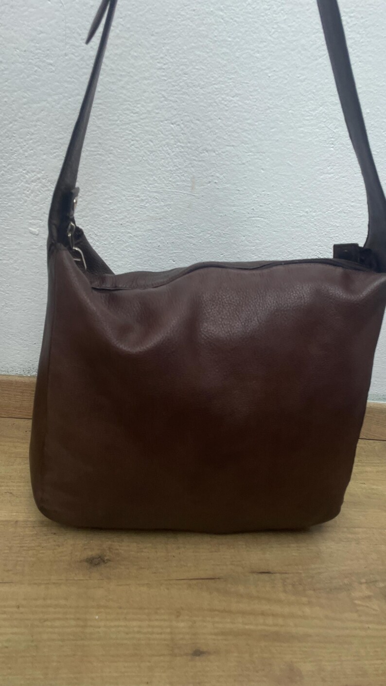 May include: A brown leather shoulder bag with a long strap. The bag is rectangular with a rounded top and a smooth, textured surface. The strap is dark brown and attached to the bag with metal hardware. The bag is sitting on a wooden surface.