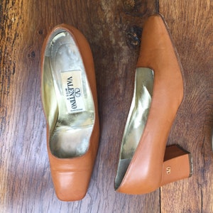 May include: A pair of tan leather Valentino Miss V heels with square toes and block heels. The interior is gold-toned, and the heels have a small gold logo. The shoes are displayed on a wooden surface.
