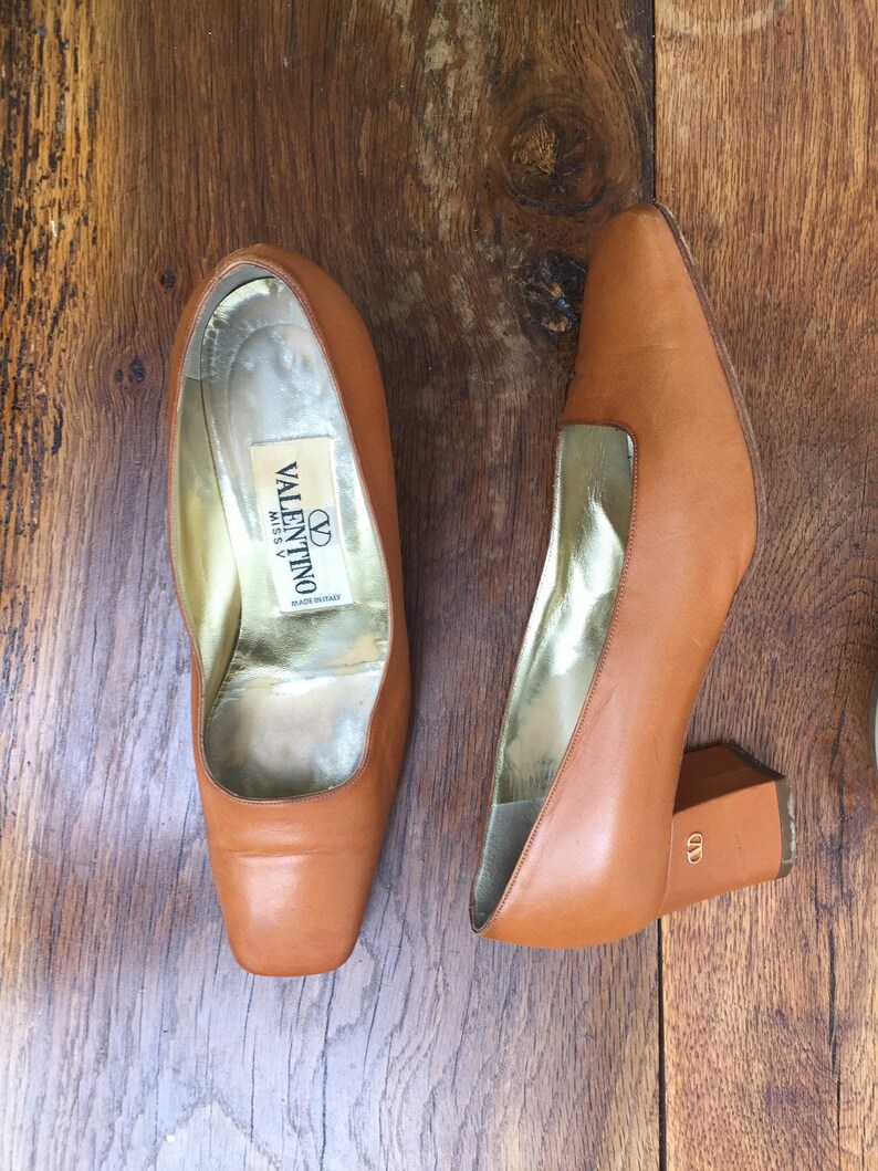 May include: A pair of tan leather Valentino Miss V heels with square toes and block heels. The interior is gold-toned, and the heels have a small gold logo. The shoes are displayed on a wooden surface.