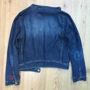 Peut inclure: Une veste en jean bleu fonc&eacute; avec un petit patch carr&eacute; au dos. La veste a un col boutonn&eacute; et des manches longues.