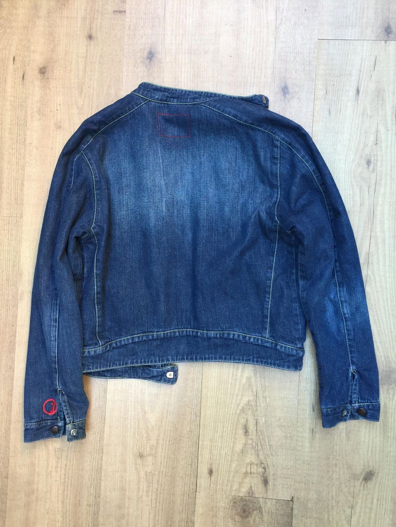 Peut inclure: Une veste en jean bleu fonc&eacute; avec un petit patch carr&eacute; au dos. La veste a un col boutonn&eacute; et des manches longues.