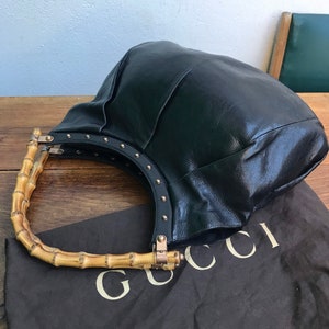 May include: Black leather handbag with bamboo handles. The bag features a rounded shape and is adorned with gold-colored studs along the handle attachments. The bag rests on a brown cloth with the brand name printed in white.