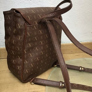 K&ouml;nnte beinhalten: Ein brauner Lederrucksack mit einem Monogrammbild. Der Rucksack hat verstellbare Tr&auml;ger und einen Klappenverschluss.