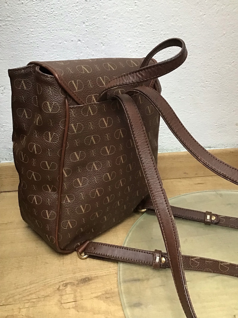 K&ouml;nnte beinhalten: Ein brauner Lederrucksack mit einem Monogrammbild. Der Rucksack hat verstellbare Tr&auml;ger und einen Klappenverschluss.