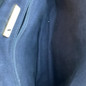 May include: Interior view of a navy blue handbag. The bag features a zippered pocket and a gold-colored rectangular accent. The lining is a solid navy blue fabric.
