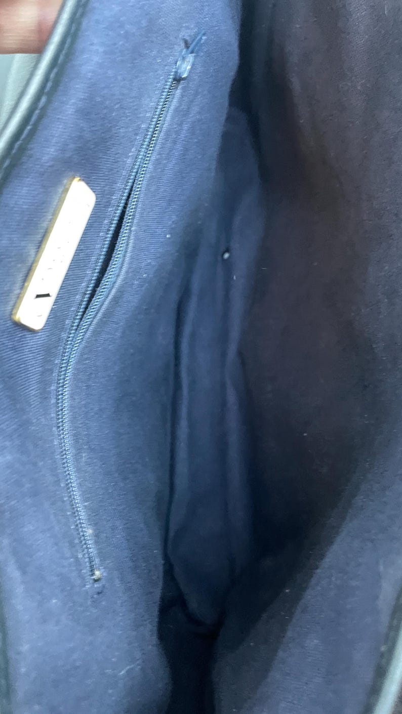 May include: Interior view of a navy blue handbag. The bag features a zippered pocket and a gold-colored rectangular accent. The lining is a solid navy blue fabric.