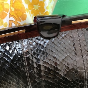 Op de afbeelding: Een vintage zwarte slangenleren clutch met een glanzende afwerking. De rechthoekige tas heeft een donkerbruin frame en een zwarte sluiting. De tas is geaccentueerd met goudkleurige metalen details en een gestikte naad in het midden.