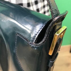 Puede incluir: Un bolso de hombro de charol en color verde azulado oscuro con acabado brillante y una hebilla de metal dorado en la correa lateral. El bolso presenta costuras visibles en los bordes y una silueta estructurada con cierre de solapa y correa.