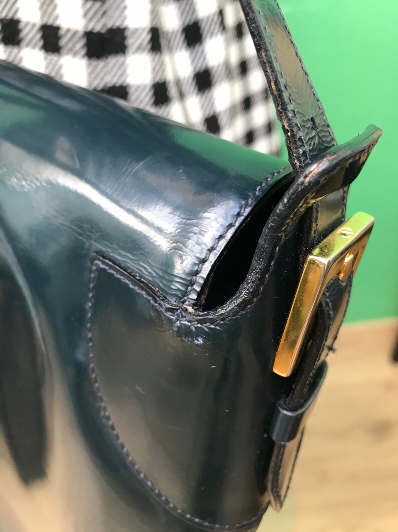 Puede incluir: Un bolso de hombro de charol en color verde azulado oscuro con acabado brillante y una hebilla de metal dorado en la correa lateral. El bolso presenta costuras visibles en los bordes y una silueta estructurada con cierre de solapa y correa.