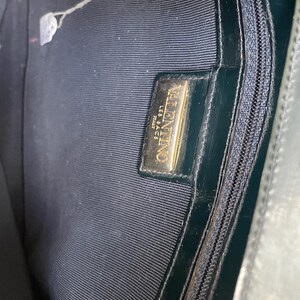 Puede incluir: El interior de un bolso de mano verde oscuro con forro de tela oscura y un bolsillo con cremallera. En el interior hay una placa rectangular de metal dorado grabada con el texto VALENTINO LES SACS ITALY. Se observa un tirador de cremallera dorado.