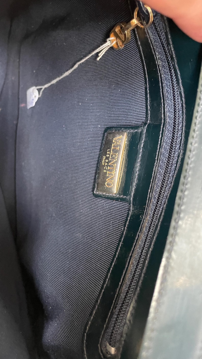 Puede incluir: El interior de un bolso de mano verde oscuro con forro de tela oscura y un bolsillo con cremallera. En el interior hay una placa rectangular de metal dorado grabada con el texto VALENTINO LES SACS ITALY. Se observa un tirador de cremallera dorado.
