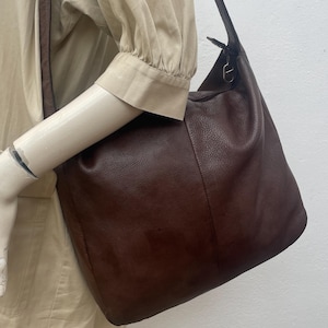 May include: A dark brown leather shoulder bag with a long strap. The bag has a soft, slouchy shape and a textured surface. The strap is attached to the bag with metal hardware.