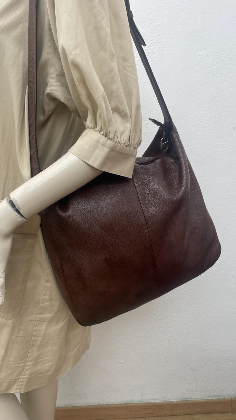 May include: A dark brown leather shoulder bag with a long strap. The bag has a soft, slouchy shape and a textured surface. The strap is attached to the bag with metal hardware.