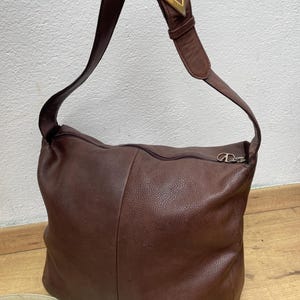 May include: A brown leather shoulder bag with a long strap. The bag has a zipper closure and a gold-colored metal buckle with a "V" design on the strap. The bag is made of a soft, textured leather.