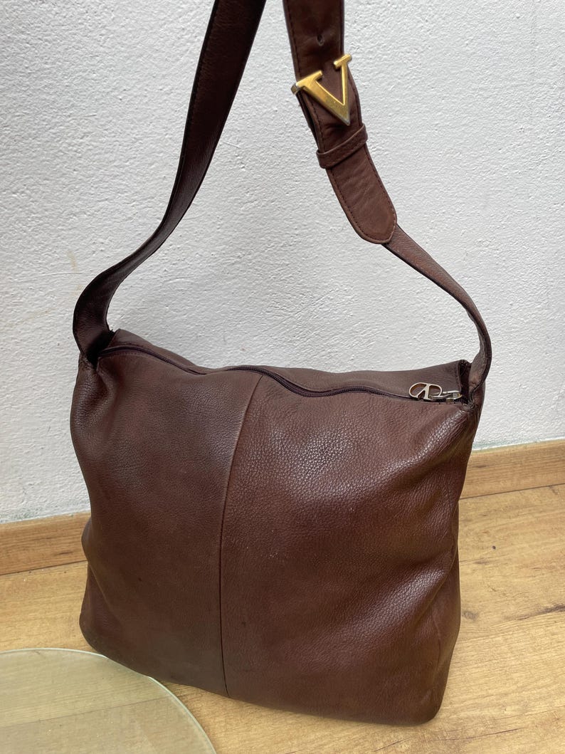 May include: A brown leather shoulder bag with a long strap. The bag has a zipper closure and a gold-colored metal buckle with a "V" design on the strap. The bag is made of a soft, textured leather.