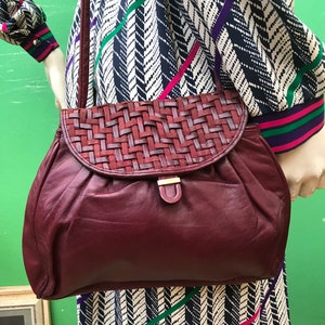 May include: A deep burgundy leather shoulder bag featuring a woven flap closure with a matching leather strap and a small gold-tone metal clasp. The main body of the bag has a soft, gathered texture and includes a long, thin shoulder strap.