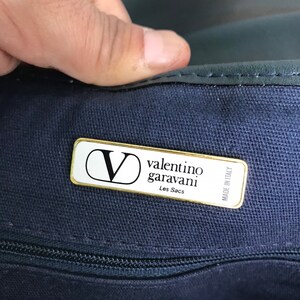 May include: Close-up of a navy blue handbag with a gold-trimmed label. The label reads "Valentino Garavani Les Sacs Made in Italy" with the brand's "V" logo. The bag's zipper is visible.