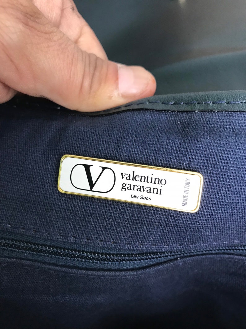 May include: Close-up of a navy blue handbag with a gold-trimmed label. The label reads "Valentino Garavani Les Sacs Made in Italy" with the brand's "V" logo. The bag's zipper is visible.