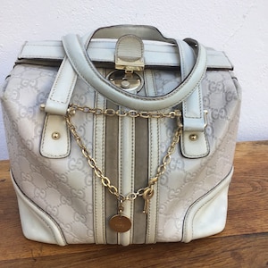 Peut inclure: Sac à main de couleur claire avec une chaîne et une clé dorées. Le sac présente une marque en relief et un design structuré. Les poignées et les bordures sont d'une teinte plus claire, et le sac est de forme rectangulaire.