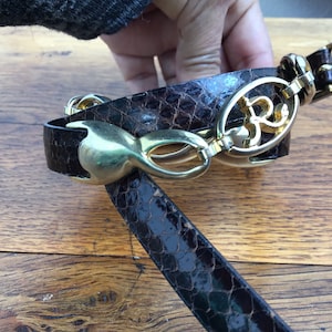 Può includere: Cintura in pelle di serpente marrone con fibbia e dettagli dorati. La cintura presenta un motivo a pelle di serpente strutturato e una fibbia dorata decorativa con un design ornato. Un accessorio elegante.