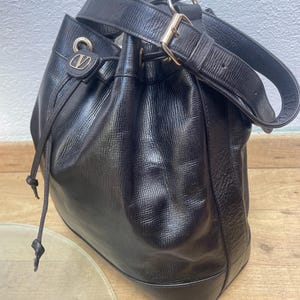 May include: Black leather bucket bag with a drawstring closure and a shoulder strap. The bag features gold-tone hardware, including a buckle on the strap and a logo detail on the front. The bag has a textured surface.