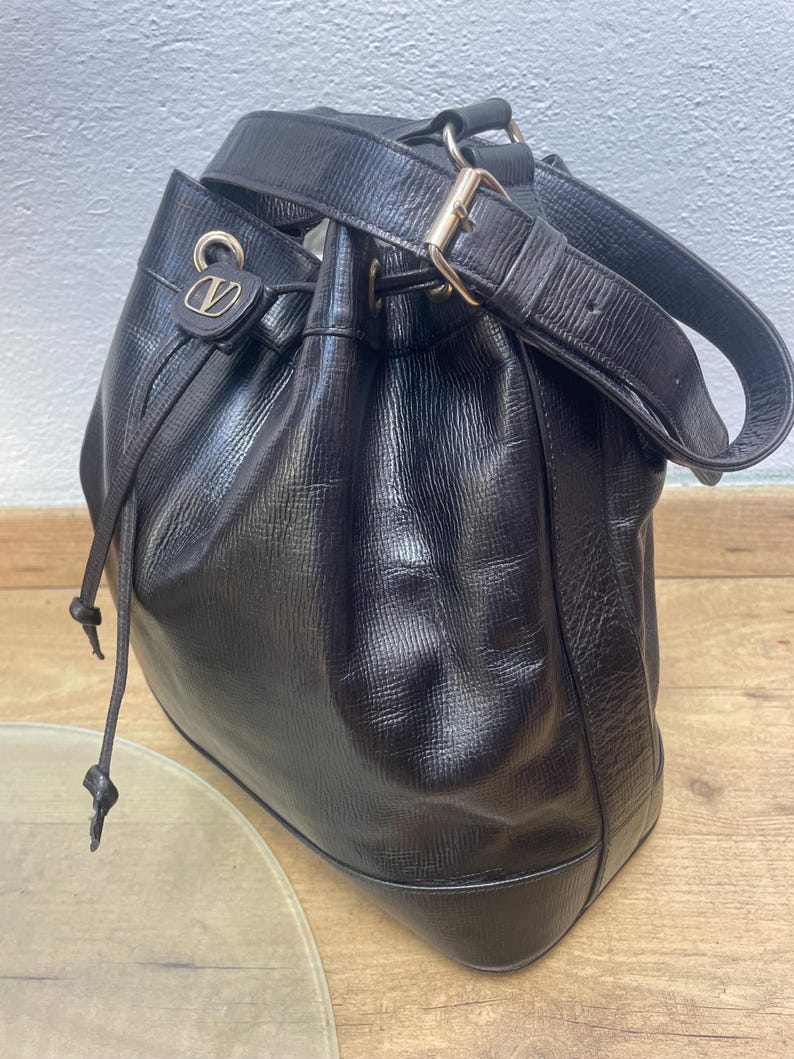 May include: Black leather bucket bag with a drawstring closure and a shoulder strap. The bag features gold-tone hardware, including a buckle on the strap and a logo detail on the front. The bag has a textured surface.