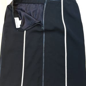 Può includere: Gonna nera con fascia in pelle e cerniera frontale. La gonna presenta due strisce bianche verticali. L'etichetta del marchio è visibile all'interno. La gonna è lunga circa 50 cm.
