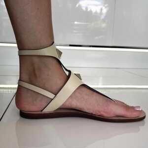May include: Cream-colored leather sandals with a T-strap design and ankle strap. The sandals feature small, gold-colored pyramid studs and a flat sole. The foot is visible, showcasing the sandal's design.