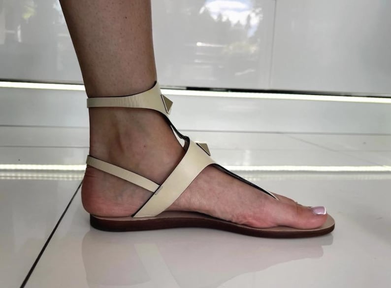 May include: Cream-colored leather sandals with a T-strap design and ankle strap. The sandals feature small, gold-colored pyramid studs and a flat sole. The foot is visible, showcasing the sandal's design.