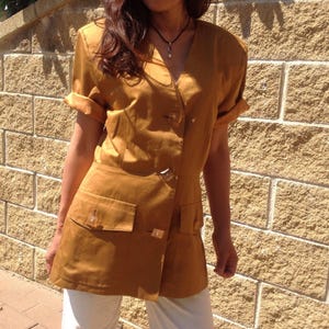 May include: Mustard-coloured short-sleeved top with a V-neck, two front pockets, and a button closure. The sleeves are rolled up. The top is paired with white trousers. The top has a tailored fit.