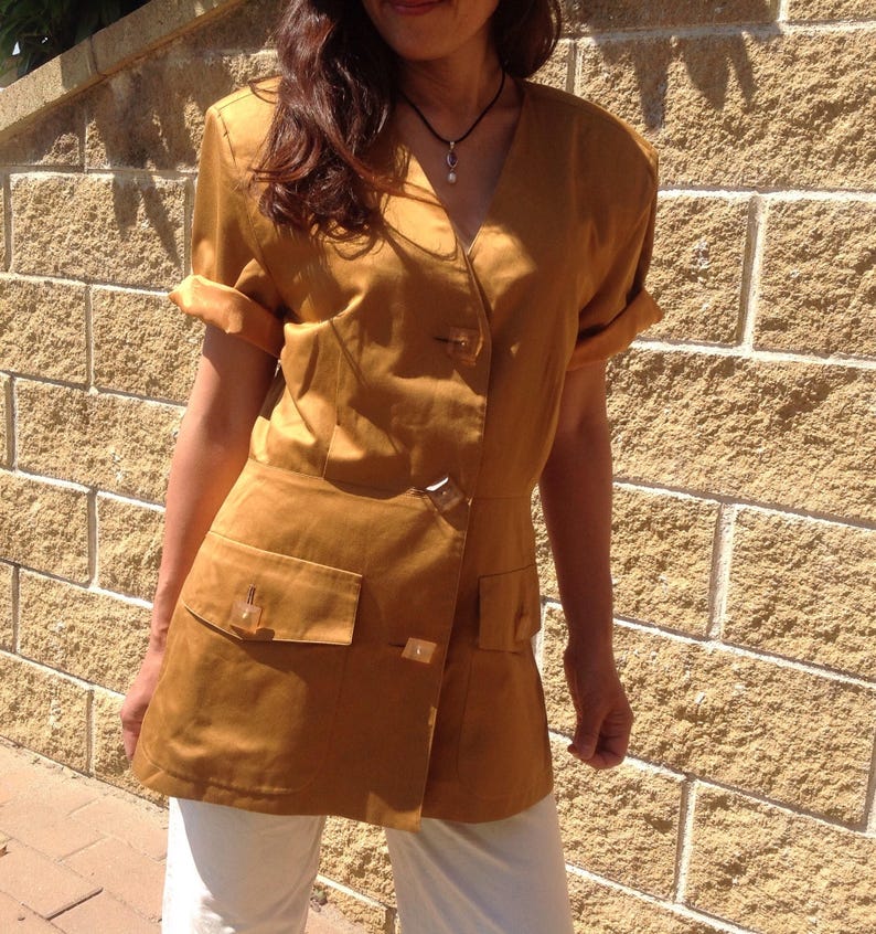 May include: Mustard-coloured short-sleeved top with a V-neck, two front pockets, and a button closure. The sleeves are rolled up. The top is paired with white trousers. The top has a tailored fit.