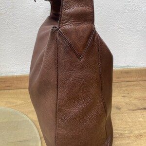 May include: A brown leather shoulder bag with a single strap and a buckle closure. The bag has a rounded shape and a textured surface. The strap is attached to the bag with a metal buckle. The bag is sitting on a wooden surface.